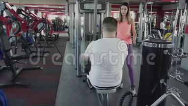 人与私人教练在健身房的训练模拟器上摇动他的手臂肌肉。 个人减肥训练